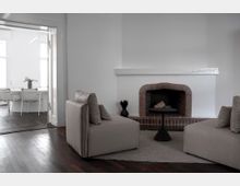 Wohnzimmer mit minimalistischem Design, weiß gestrichenen Wänden und einem gemauerten Kamin mit dunkler Öffnung. Im Vordergrund stehen zwei beige gepolsterte Sessel und ein kleiner schwarzer Beistelltisch; im Hintergrund ist ein angrenzender Raum mit einem weißen Tisch und Stühlen sichtbar.