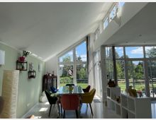 Ein modernes Wohnzimmer mit hohen, schrägen Decken und großen Fenstern, die den Raum mit Tageslicht fluten. Der Raum ist mit bunten Stühlen um einen Glastisch, einer grünen Wand mit Dekoelementen und einer offenen Sicht auf einen grünen Garten eingerichtet.