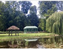 Pavillon aus grünem Metall mit filigranen Verzierungen inmitten eines Parks, umgeben von grüner Wiese, Bäumen und einer Weide am rechten Bildrand; im Vordergrund ein kleiner Teich mit Schilfpflanzen.
