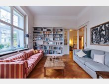 Wohnzimmer mit einem großen Fenster links, einer weißen Bücherwand in der Mitte und einer Schiebetür zum angrenzenden Raum im Hintergrund. Im Vordergrund stehen ein gestreiftes Sofa und ein graues Sofa gegenüber, getrennt durch einen Holztisch auf Parkettboden.