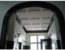 Blick in einen Flur mit weißem Kassettendeckenmuster, dunklen Holzabschlüssen und Wandleuchten. Am Ende des Flurs befindet sich ein großes Fenster, das Tageslicht hereinlässt.