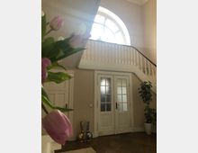 Ein helles, klassisches Treppenhaus mit einem großen, halbkreisförmigen Fenster über einer weißen Holzbalustrade. Im Vordergrund sind rosa Tulpen zu sehen, während im Hintergrund eine Doppeltür mit Milchglas und Dekoration wie Figuren und eine Zimmerpflanze das Ambiente prägen.