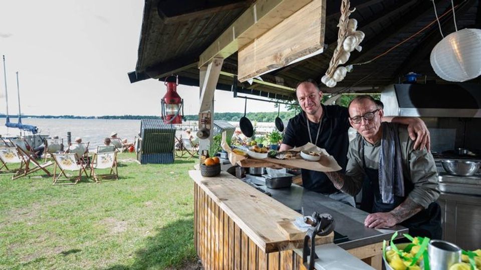 Eine offene Strandbar aus Holz mit Blick auf einen See. Im Vordergrund befindet sich eine Theke mit Kochutensilien, im Hintergrund sind Liegestühle, Sonnenschirme und Boote am Ufer zu sehen.