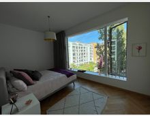 Schlafzimmer mit minimalistischem Design, einem Bett mit rosa und violetten Kissen, einem Nachttisch und einem großen Fenster, das den Blick auf moderne Wohngebäude und Bäume draußen freigibt. Der Raum hat helle Wände, einen Holzboden und einen Teppich mit geometrischem Muster.