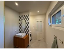 Modernes Badezimmer mit hellem Design, einer Waschbeckeneinheit aus Holz, ovalem Spiegel und gemusterter Fliesenrückwand. Rechts an der Wand befindet sich ein Fenster und darunter Handtuchhaken, im Hintergrund eine weiße Tür und eine Waschmaschine.