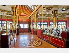 Innenraum eines asiatischen Restaurants mit opulenter Dekoration, roten Wänden und goldenen Säulen. Die Decke ist reich verziert und von kleinen Lichtern beleuchtet, während Glasscheiben mit floralen Mustern und runde Fenster den Raum schmücken.