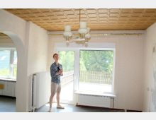 Ein einfacher Wohnraum mit cremefarbenen Wänden, einem goldfarbenen Kassettendesign an der Decke und einer zentralen Hängeleuchte. Der Raum hat große Fenster mit Blick auf einen Gartenzaun und Bäume sowie Heizkörper unter den Fenstern.