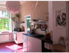 Helle Küche mit rotem Boden, schwarzer Spüle und weißen Schränken. Links ein geöffnetes Fenster mit Blick auf eine begrünte Fassade, während Regale und eine Hängelampe den Raum dekorieren.