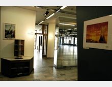 Innenraum eines öffentlichen Gebäudes mit Fliesenboden und weißen Wänden. Auf der linken Seite ein kleiner Holztisch mit Prospekthaltern, daneben Schilder mit der Aufschrift „WC Herren“. Rechts ein Spiegel und ein gerahmtes Bild, im Hintergrund ein Bereich mit großen Fenstern und Tageslichteinfall.