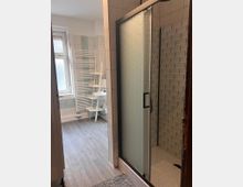 Badezimmer mit gefliester Dusche und mattierter Glas-Schiebetür, daneben eine strukturierte Wandverkleidung im Duschbereich. Im Hintergrund stehen ein Waschbecken, ein weißer Handtuchheizkörper und ein Fenster; der Boden ist in grauer Holzoptik gehalten.