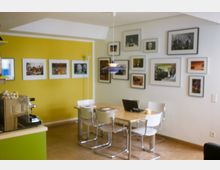 Ein kleiner, moderner Innenraum mit einer gelben Akzentwand, an der Fotografien in unterschiedlichen Größen hängen. Ein Holztisch mit vier weißen Stühlen steht im Vordergrund, daneben eine Kaffeestation und eine Deckenleuchte über dem Tisch.