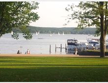 Park mit Blick auf einen See, im Vordergrund eine Grünfläche und Spaziergänger, im Hintergrund Segelboote auf dem Wasser und an einem Anleger vertäute Motorboote. Umgeben von Bäumen und Wald am gegenüberliegenden Ufer.