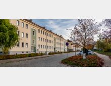 Wohnblock mit drei Stockwerken in heller Putzfassade, teilweise grün abgesetzt, entlang einer gepflasterten Straße mit Gehwegen. Im Vordergrund ein Baum mit herbstlichem Laub sowie Straßenschilder und parkende Autos.