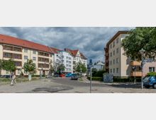 Eine Wohnstraße mit mehrstöckigen Wohngebäuden im klassischen Stil, cremefarbene Fassaden mit roten Ziegeldächern und einigen Balkonen. Die Szene zeigt geparkte Autos, Fußwege, kleine Bäume und eine Beschilderung, unter grauen, bewölkten Himmel.