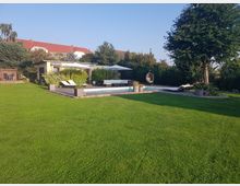 Großer Garten mit einem Poolbereich, umgeben von gepflegtem Rasen und Hecken. Der Poolbereich enthält Liegestühle, Sonnenschirme, Pflanzenkübel, eine Sitzecke und eine kleine Gartenhütte.