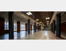 Großer Innenraum mit Marmorboden, schwarzen Säulen mit roten Akzenten und einer Holzdecke mit geometrischem Muster. Im Hintergrund befindet sich eine breite Treppe, während die Fensterfronten an der Seite Tageslicht einlassen.