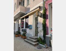 Fassade eines Stadtgebäudes mit cremefarbener Putzwand und gelb-weiß gestreifter Markise über einer Tür. Vor dem Eingang befinden sich Treppenstufen, Topfpflanzen und Kletterpflanzen an der Wand, daneben ein kleines Schild mit Aufschrift.