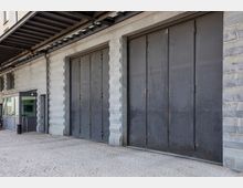 Außenansicht eines Gebäudes mit grauen Metalltoren und massiven, geometrisch gemusterten Steinsäulen. Links befindet sich ein kleiner verglaster Eingangsbereich, der von einer Überdachung aus Stahl geschützt wird.