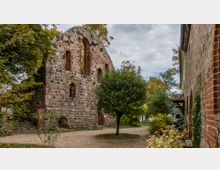 Ruinen eines historischen Gebäudes aus unregelmäßigem Feldstein und roten Ziegeln, umgeben von Bäumen und Pflanzen. Der Boden ist von einem Weg durchzogen, und im Hintergrund ist ein See sichtbar.