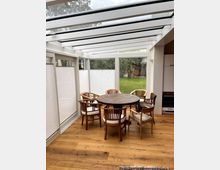 Ein Wintergarten mit gläsernem Dach und weißen Rahmen, ausgestattet mit einem runden Holztisch und mehreren Holzstühlen. Große Fenster erlauben einen Blick auf einen Garten mit Rasen, Bäumen und einer Holzhütte im Hintergrund.