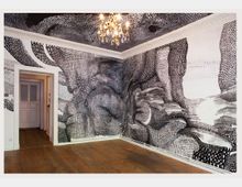 Ein Zimmer mit Holzboden, verzierten weißen Türrahmen und Wänden, die mit abstrakten, schwarzen Zeichnungen auf weißem Hintergrund dekoriert sind; an der Decke hängt ein Kronleuchter.