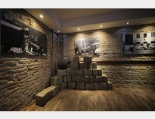 Ausstellungsraum mit einer Backsteinwand, an der historische Schwarz-Weiß-Fotografien hängen. Im Vordergrund ist eine Pyramide aus Pflastersteinen auf einem Holzboden zu sehen, beleuchtet von Deckenstrahlern.