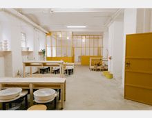 Ein heller Raum mit weiß gestrichenen Backsteinwänden und gelben Türen und Fensterrahmen. Im Vordergrund stehen Holztische und Töpferscheiben, im Hintergrund befindet sich ein abgetrennter Bereich durch gelbe Glastrennwände.