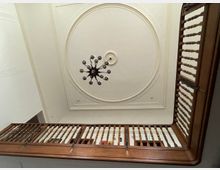 Blick von unten durch ein Treppenhaus mit Holzgeländern, das zu einer Decke mit einem großen, runden Stuckornament und einem schwarzen Kronleuchter führt.