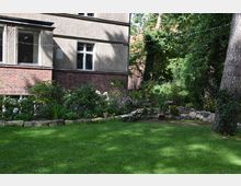 Grüne Rasenfläche mit einem bepflanzten Gartenbereich im Vordergrund, eingefasst von einer Steinkante; dahinter ein Wohngebäude mit Fassaden aus Putz und Ziegelstein sowie große Fenster. Im Hintergrund sind Bäume und dichtes Grün zu sehen.