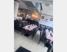 Innenansicht eines Restaurants mit modernem Design. Mehrere Tische sind mit rot-weiß karierten Tischdecken, gefalteten Servietten und kleinen Pflanzen dekoriert, während im Hintergrund eine Bar mit beleuchteten Regalen und Getränkeflaschen zu sehen ist.