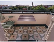 Kleiner Balkon mit gemustertem Fliesenboden, einem klappbaren Holztisch und einem grünen Klappstuhl. Im Hintergrund eine Stadtansicht mit Bäumen, Wohngebäuden und der Turmspitze einer Kirche.