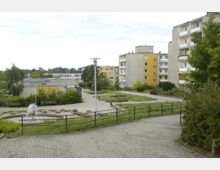 Wohngebiet mit mehrstöckigen Plattenbauten, teils in gelber Fassade. Im Vordergrund ein kleiner Park mit einem Brunnen, Bänken und Grünanlagen, umgeben von gepflasterten Wegen und vereinzelten Bäumen.