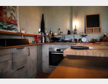 Moderne Küche mit hellem Holz und grauen Fronten, ausgestattet mit einem schwarzen Spülbecken, einer Arbeitsplatte mit Küchenutensilien, einem Herd mit Backofen und einer Küchenwaage. Im Hintergrund befinden sich ein schlichtes Wandregal mit Gewürzen, Flaschen und einer kleinen Tafel.