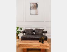 Wohnzimmer mit einer schwarzen Ledercouch vor einer weißen Wand mit dezentem Wandleisten-Dekor. Ein abstraktes Gemälde hängt an der Wand, davor ein rechteckiger Couchtisch aus Holz mit drei schwarzen, dekorativen Kerzenhaltern; links im Hintergrund eine Zimmerpflanze in goldfarbenen Übertöpfen.