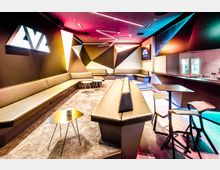 Modern gestalteter Lounge-Bereich mit geometrischen Wandmotiven, langen gepolsterten Sitzbänken, kleinen Tischen und einem Tresen. Die Beleuchtung ist in bunten Farbtönen gestaltet, und es gibt einen Kühlschrank mit Getränken im Hintergrund.