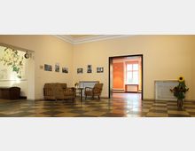 Ein Innenraum mit beigen Wänden und einem Schachbrettboden aus Fliesen. Im Vordergrund stehen zwei gepolsterte Stühle und ein kleiner Tisch, an den Wänden hängen Schwarz-Weiß-Bilder. Im Hintergrund führt eine offene Tür zu einem weiteren Raum mit einem großen Fenster, das Tageslicht hereinlässt.