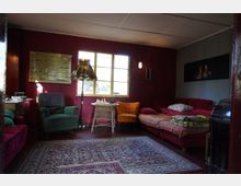 Gemütliches Wohnzimmer mit roten Wänden, einem großen Teppich am Holzboden und verschiedenen Sitzmöbeln, darunter ein grüner und ein orangefarbener Sessel sowie eine rote Couch. An der Wand hängen ein Bild mit Schiffsmotiven und ein Kaminbild, ein Fenster lässt Tageslicht herein.