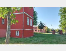 Roter Backsteinbau mit weißen Fensterrahmen in einer parkähnlichen Umgebung, umgeben von Rasen, Bäumen und einer Ziegelmauer mit Torpfosten. Im Hintergrund weitere Gebäude und Laternen, bei klarem Himmel.