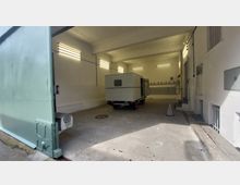 Innenraum einer einfachen Garage mit weißen Wänden und mehreren leuchtenden Neonröhren, in der ein kleiner Anhänger mit geschlossener, grauer Außenverkleidung geparkt ist. Links ist ein großes, mintgrünes Garagentor zu sehen, der Boden besteht aus Beton.