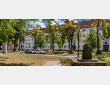 Wohngebäude mit weißer Fassade und roten Ziegeldächern im Hintergrund, umgeben von hohen Bäumen. Im Vordergrund ein kleiner Platz mit Wiese, Gehwegplatten und einem steinernen Denkmal.