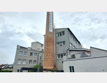 Backstein-Schornstein mit der Aufschrift „Luckenwalde“ vor einem grauen mehrstöckigen Gebäude. Im Vordergrund sind kleine Grünflächen und eine teilweise überdachte Struktur sichtbar, der Himmel ist bewölkt.