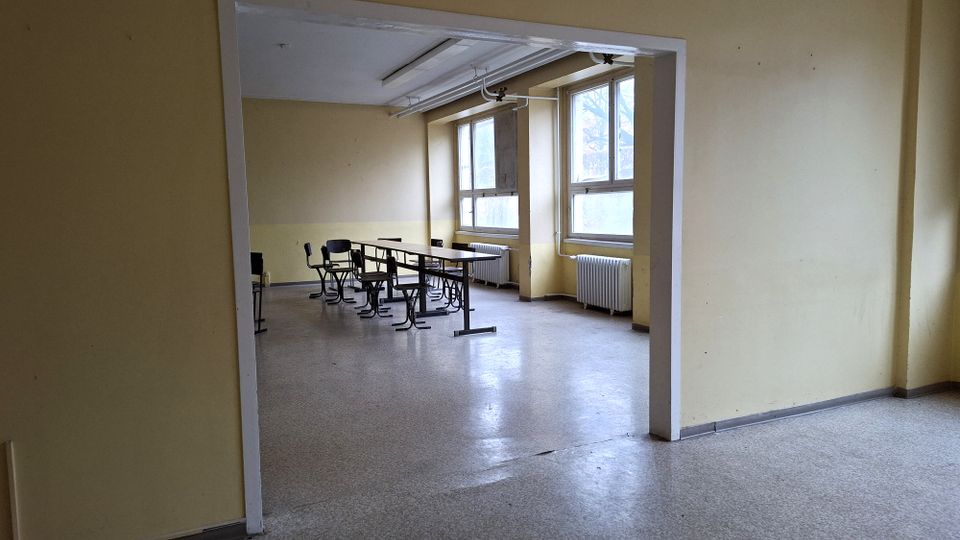 Blick durch einen breiten Durchgang in einen einfachen, leeren Klassenraum mit hellen Wänden, Linoleumboden und mehreren Schulbänken in der Mitte. Große Fenster mit Heizkörpern darunter lassen Tageslicht in den Raum; an der Decke sind lange Leuchtstofflampen montiert.