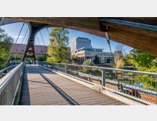 Moderne Fußgängerbrücke aus Holz und Stahl führt über einen Fluss zu einem Gebäudeensemble mit großen Glasfassaden und flachem Dach, umgeben von Bäumen und Grünflächen. Im Hintergrund steht ein mehrstöckiges Bürogebäude.