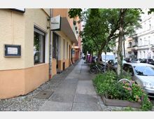 Straßenszene in einer Wohngegend mit bepflanzten Grünflächen neben einem Gehweg, geparkten Autos und Fahrrädern. Die Gebäude haben bunte Fassaden, und Geschäftsschilder sind unter einem Baum an der Hauswand sichtbar.