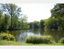 Ein ruhiger See inmitten eines Parks, umgeben von dichtem, grünem Wald. Im Vordergrund blühende Pflanzen und eine Grasfläche, während auf dem Wasser kleine Boote mit Menschen zu sehen sind.