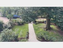 Gartenansicht mit einer gepflasterten Gehwegmitte, die von Rasen und Sträuchern gesäumt ist. Ein großer Baum steht rechts, und im Hintergrund ist ein geparktes Auto sowie eine Straße sichtbar.