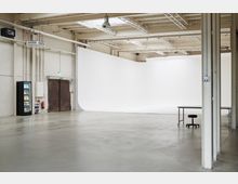 Large industrial studio space with a white cyclorama backdrop, concrete floor, and exposed ceiling beams with skylights and strip lighting. Along one wall are metal double doors, a vending machine, and visible pipes, with a table and stool in the foreground.