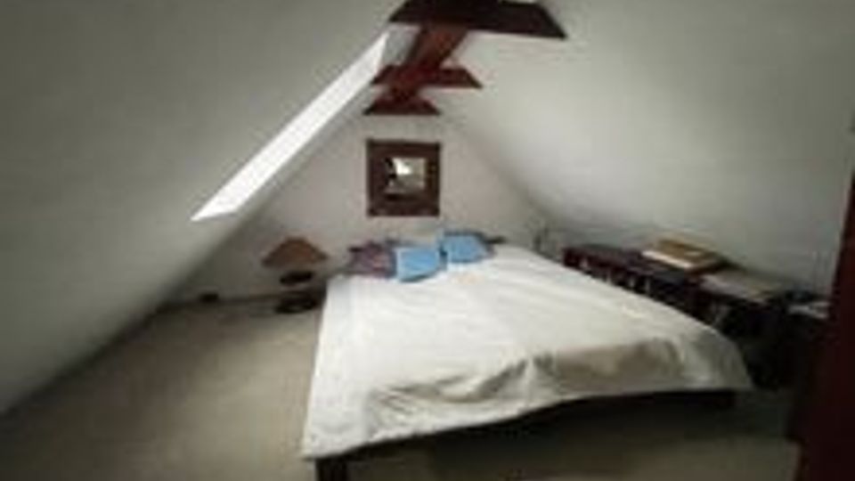 Schlafzimmer im Dachgeschoss mit schrägen weißen Wänden und sichtbaren Holzbalken, in der Mitte ein Doppelbett auf Teppichboden. An den Seiten stehen niedrige Möbel und Gegenstände, an der Stirnwand ein kleines Fenster.