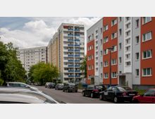 Eine Wohnstraße mit mehrstöckigen Wohngebäuden im Hintergrund, darunter ein orange-weißes Gebäude und ein Hochhaus mit Balkonen in Blau- und Beigetönen. Geparkte Autos und einige Bäume säumen die Straße, bei bewölktem Himmel.