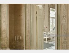 Detailreiche Innenaufnahme eines historischen Gebäudes mit verzierten Türrahmen in antikem Stil. Im Vordergrund steht ein silberner Kerzenständer, und durch eine geöffnete Tür ist ein Raum mit einer weißen Holzbank und großen Fenstern zu sehen.